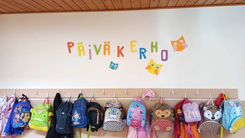 Päiväkerholaisten reppuja naulakossa, yläpuolella askarreltu teksti Päiväkerho.